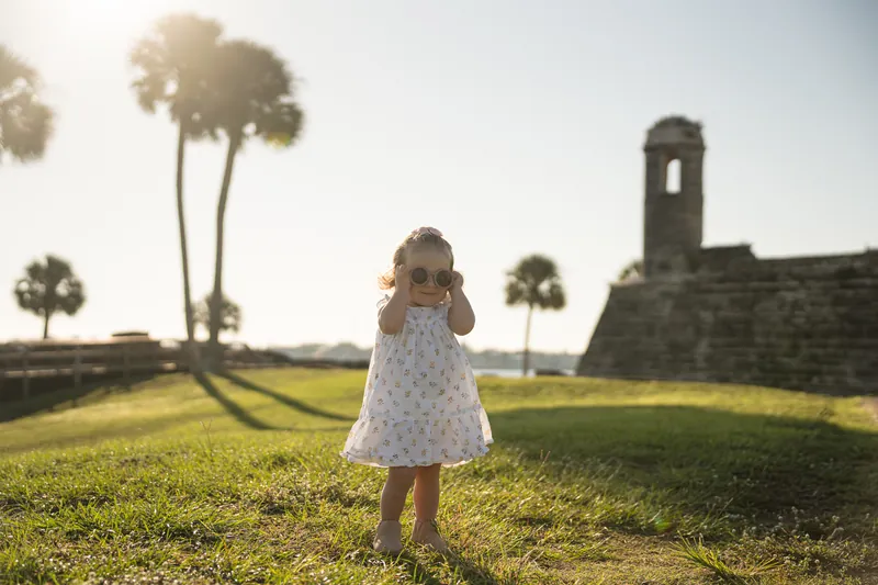A small child in a white polka dot dress stands alone on a sunny lawn with palm trees and stone ruins visible across the field.