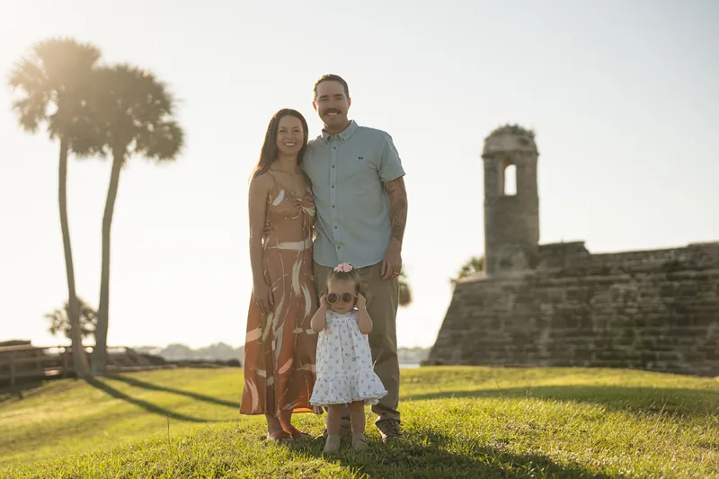 Brandon and Mallory stand with their young daughter in a white dress on green grass, with palm trees and historic fort walls behind them.