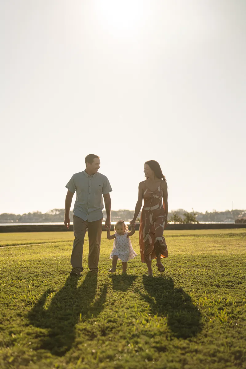 Brandon, Mallory, and Jane walk together across an open green field holding hands, casting long shadows in the golden sunlight.