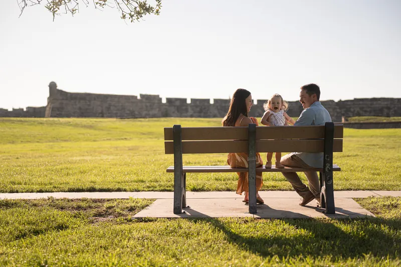 Brandon and Mallory sit on a wooden bench with Jane between them, facing the historic fort ruins across the grassy landscape.