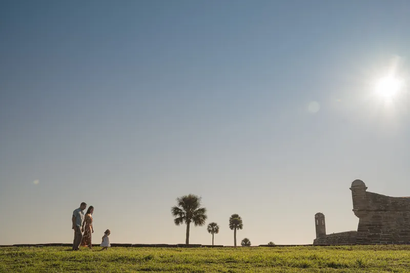 Mallory, Brandon, and Jane stand in a grassy field with palm trees and historic fort ruins under a bright Florida sky.