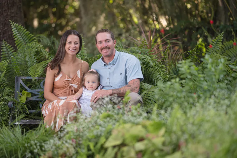 Mallory and Brandon sit together on a bench surrounded by lush green ferns, holding Jane between them in a garden setting.
