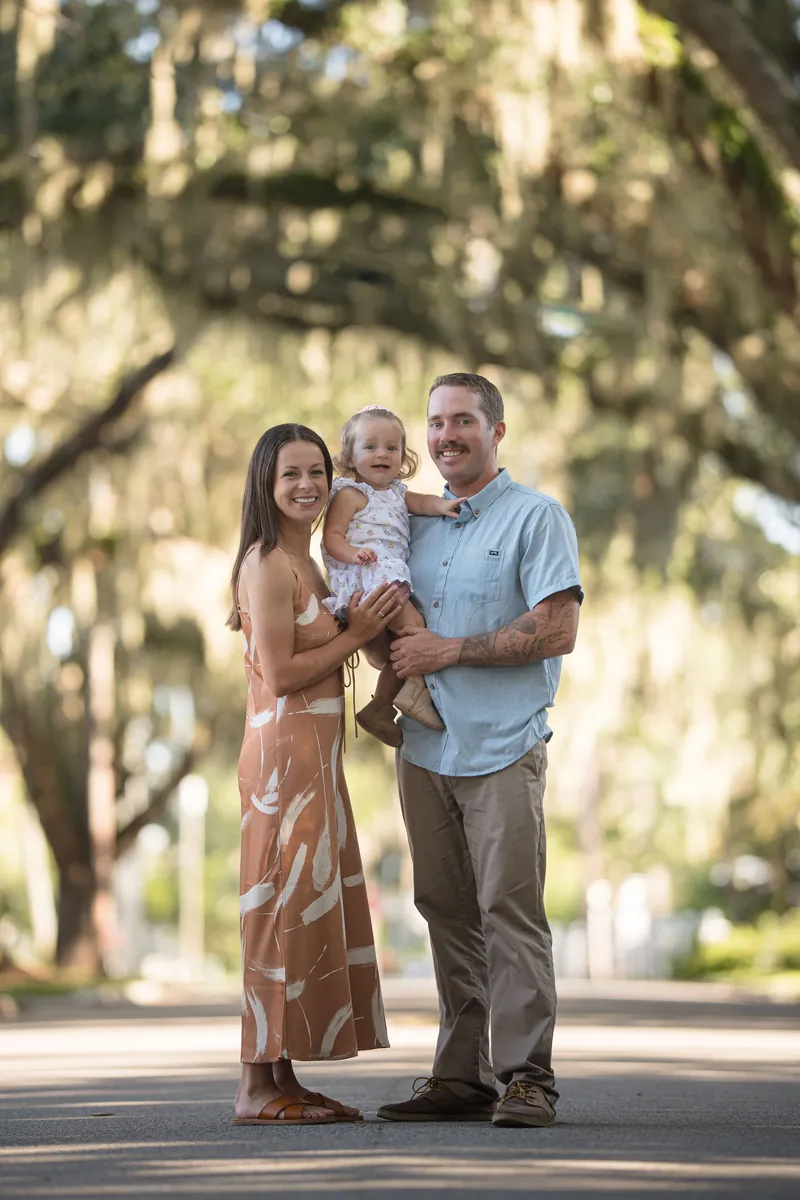 Brandon holds Jane while Mallory stands beside them under an avenue of Spanish moss-draped oak trees on a sunny afternoon.