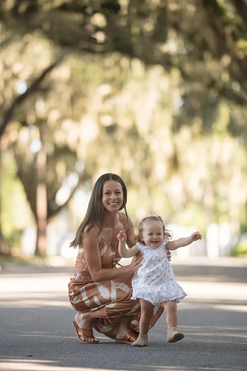 Mallory crouches down to Jane's level on a shaded tree-lined path, with Jane reaching out toward the camera with a playful expression.