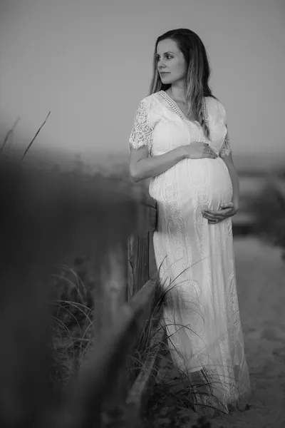Black and white portrait of expectant mother standing by beach fence, looking into the distance while cradling bump.
