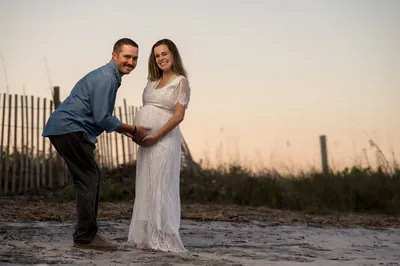Fun maternity portrait with dad leaning down to touch baby bump while both smile at camera, beach dunes behind them.