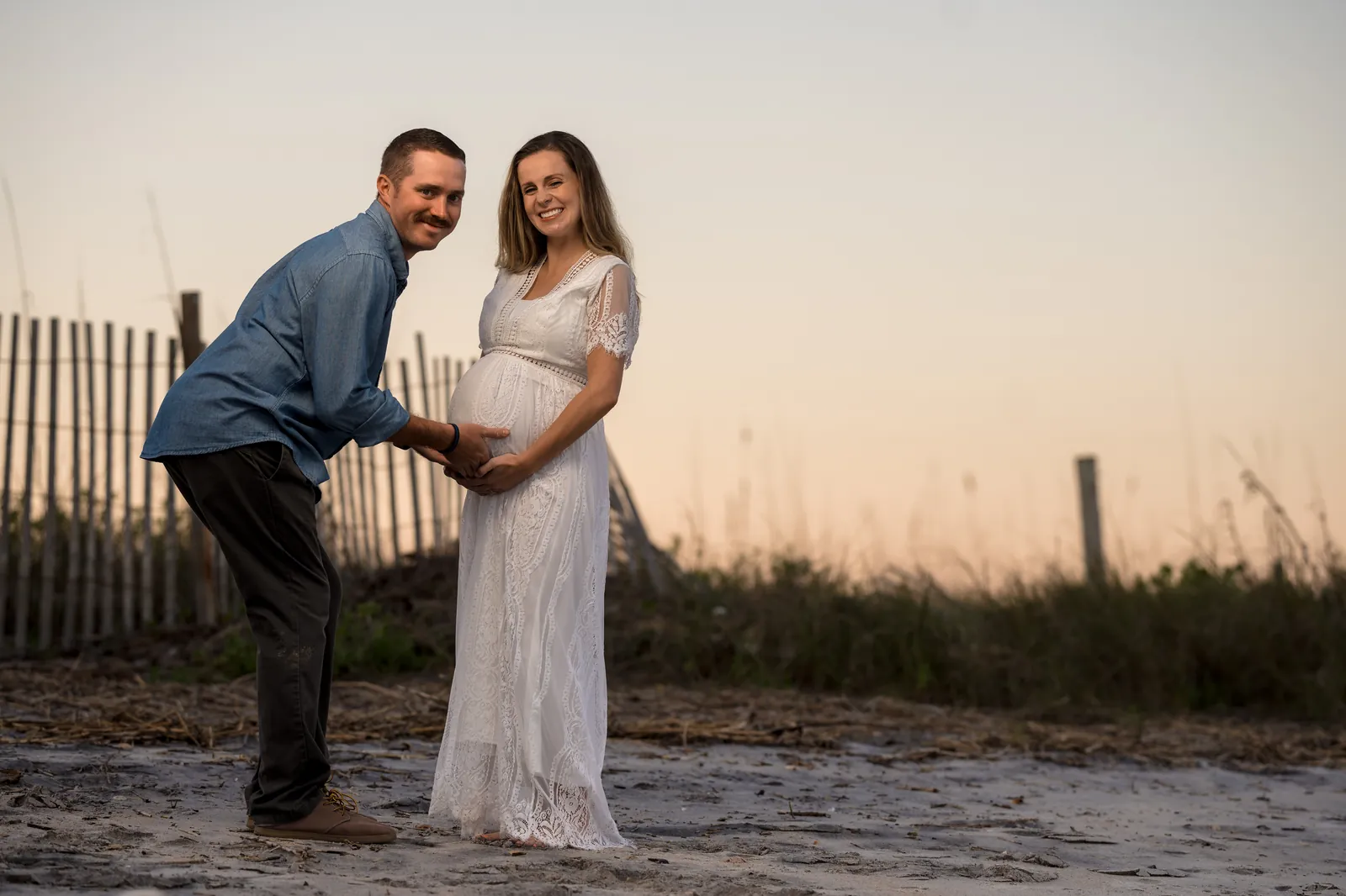 Fun maternity portrait with dad leaning down to touch baby bump while both smile at camera, beach dunes behind them.