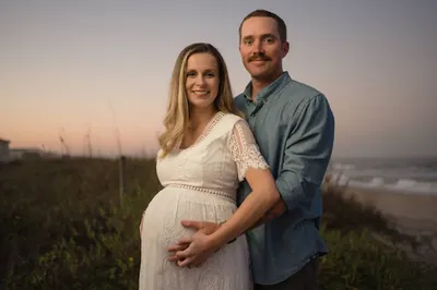 Expectant couple smiling at camera with beach dunes and ocean behind them during maternity session.