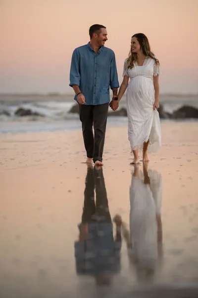 Couple walking hand in hand on wet sand with their reflection visible, pink sunset sky and jetty rocks in background.