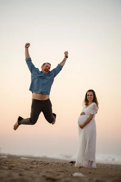 Excited dad-to-be jumping in the air while expectant mother stands smiling with hand on bump at Vilano Beach sunset.