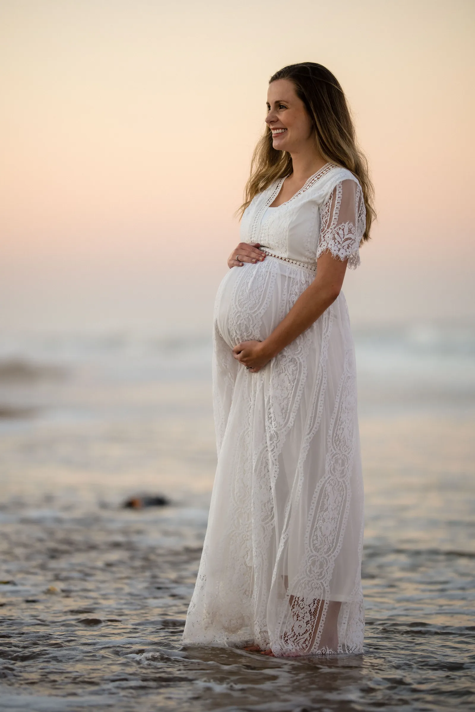 Expectant mother standing in shallow water at sunset, smiling and cradling baby bump in white lace dress.