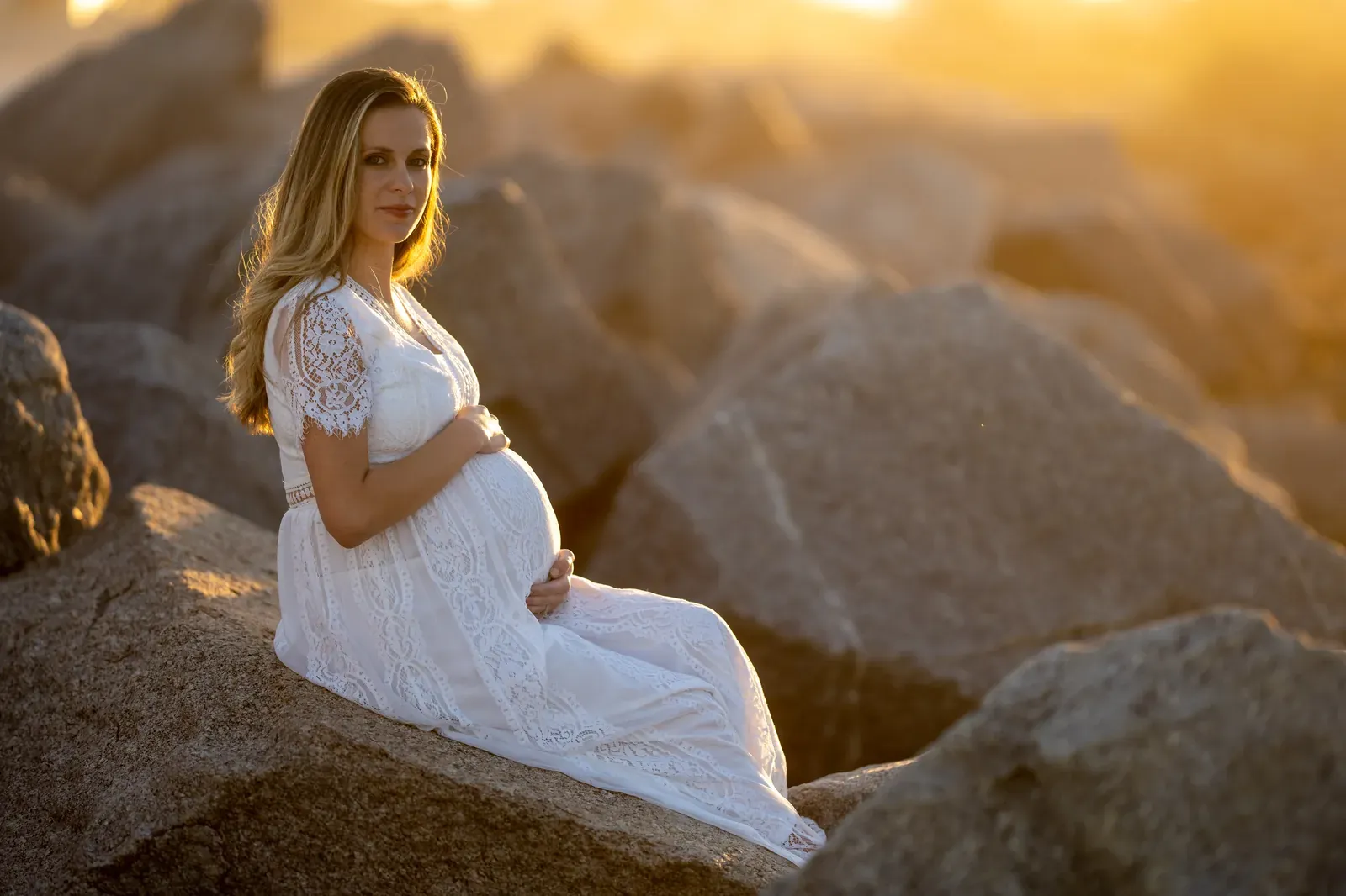 Expectant mother in white lace dress sits on jetty rocks at Vilano Beach during golden hour, cradling her baby bump with warm sunset light behind her.