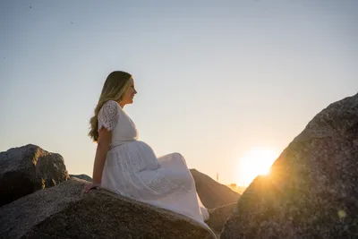 Pregnant woman in white lace dress posed on rocks at sunset