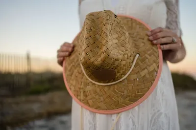 Detail shot of expectant mother holding straw hat in front of baby bump with beach fence in background.