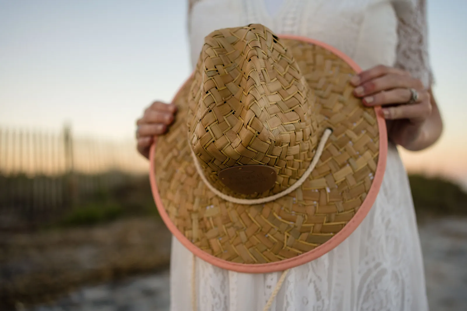 Detail shot of expectant mother holding straw hat in front of baby bump with beach fence in background.