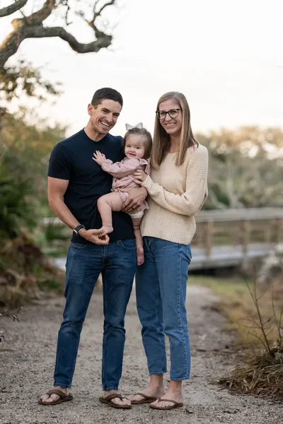 A happy family of three poses together outdoors, with dad holding their adorable baby while mom stands close beside them, all smiling warmly at the camera.