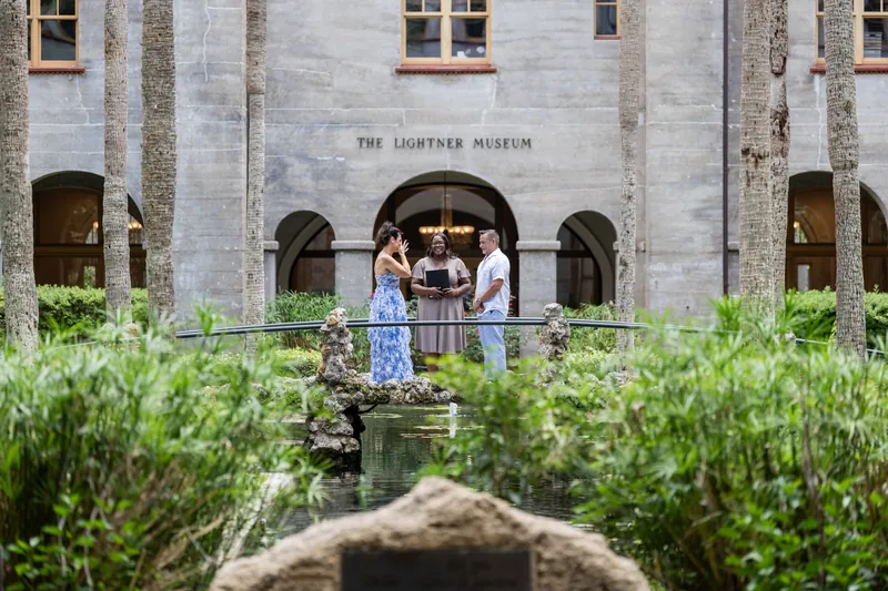 Mary and Brian stand holding hands on a railing at The Lightner Museum's courtyard, framed by arched stone architecture.