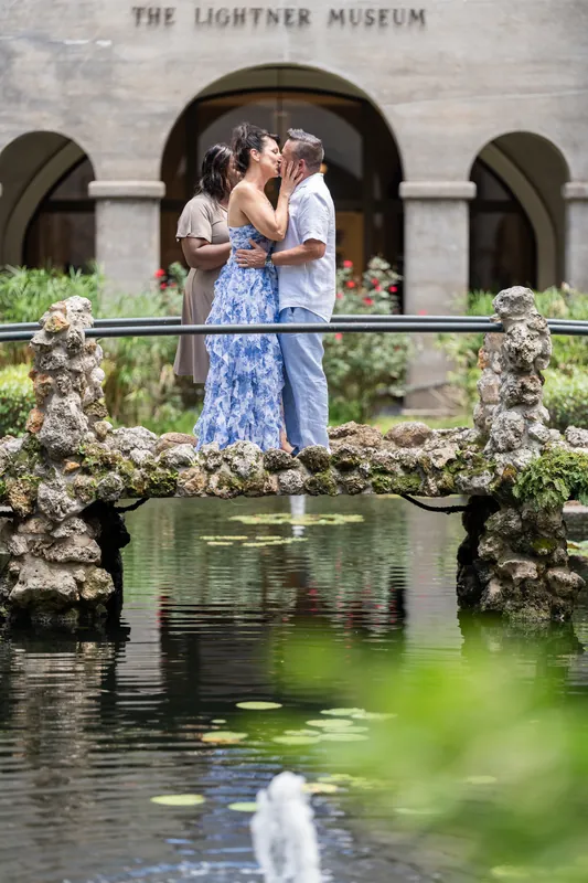 Mary and Brian kiss on a moss-covered stone bridge at The Lightner Museum, the building's arches framing them.
