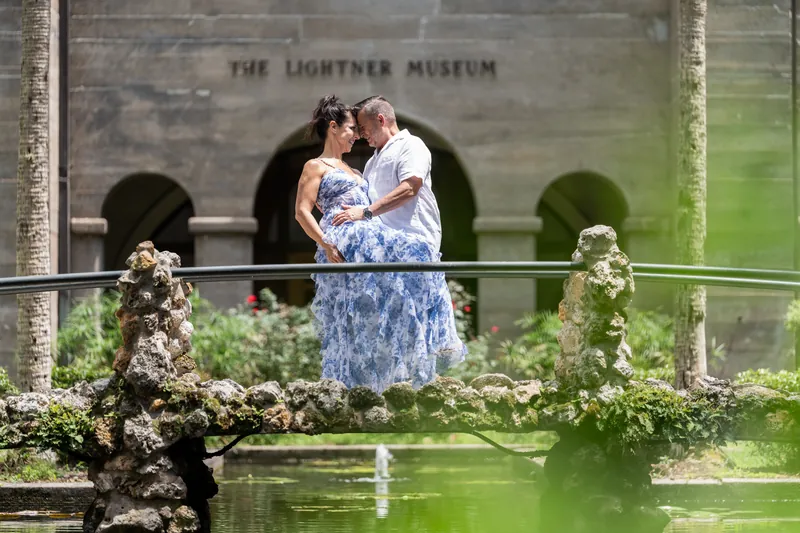 Mary and Brian embrace on a balcony overlooking a pond at The Lightner Museum, Mary in a blue floral dress.