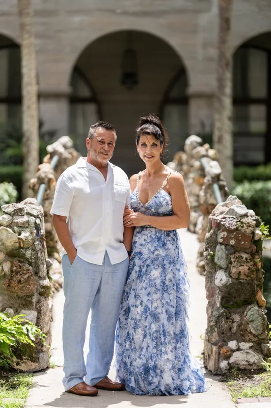 Brian and Mary stand together in the museum's courtyard surrounded by white flowering plants, both smiling.