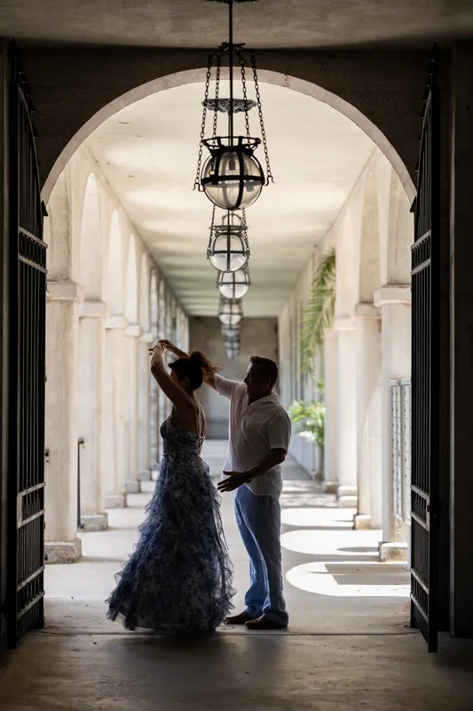 Mary in a navy ruched gown and Brian in a blue suit kiss under a hanging lantern in an arched colonnade.