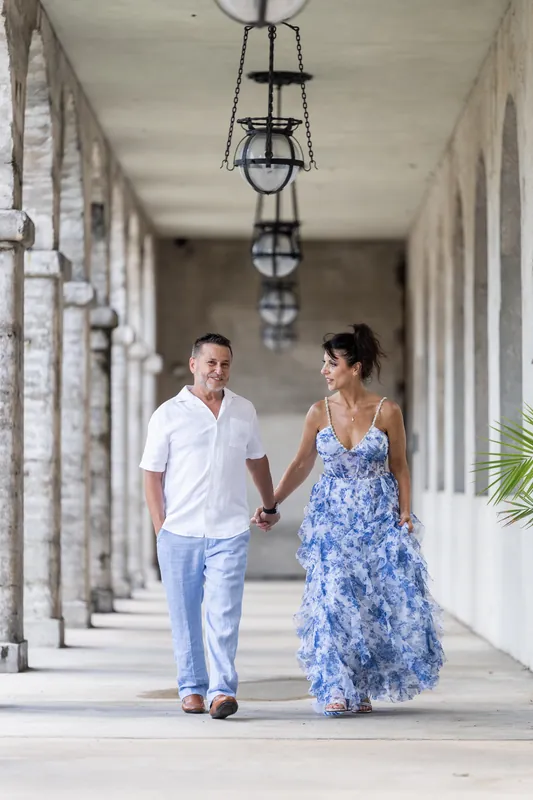 Brian holds Mary's hand while walking down a covered colonnade with hanging lanterns overhead.