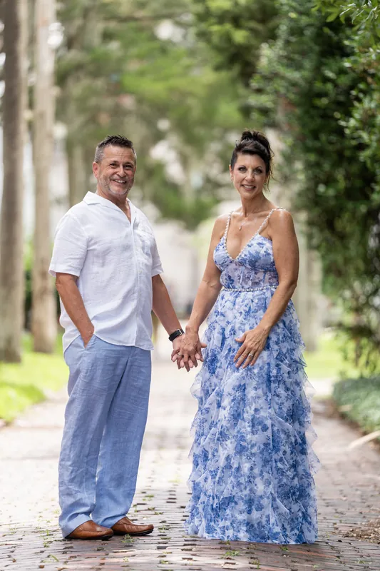 Mary and Brian hold hands while walking down a tree-lined brick pathway in dappled sunlight.