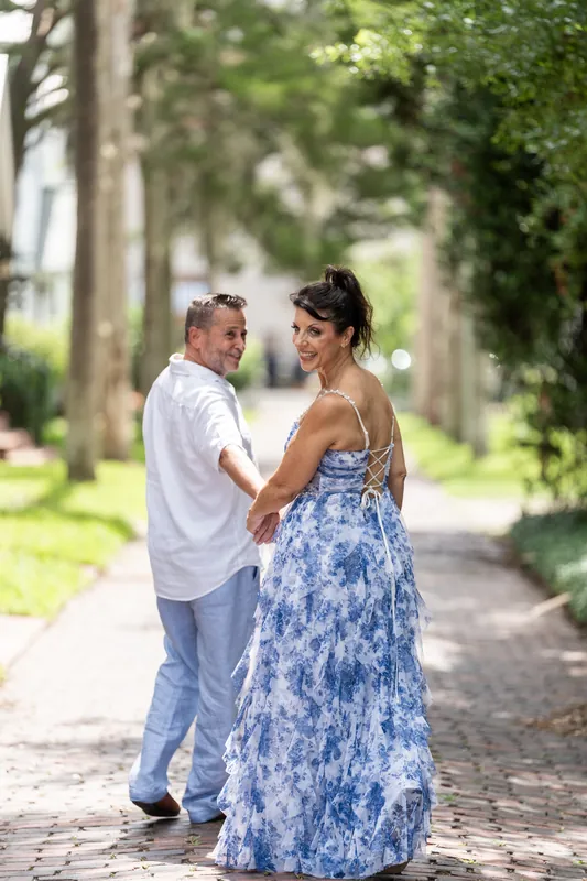 Brian and Mary face each other on a shaded brick walkway lined with green foliage on both sides.