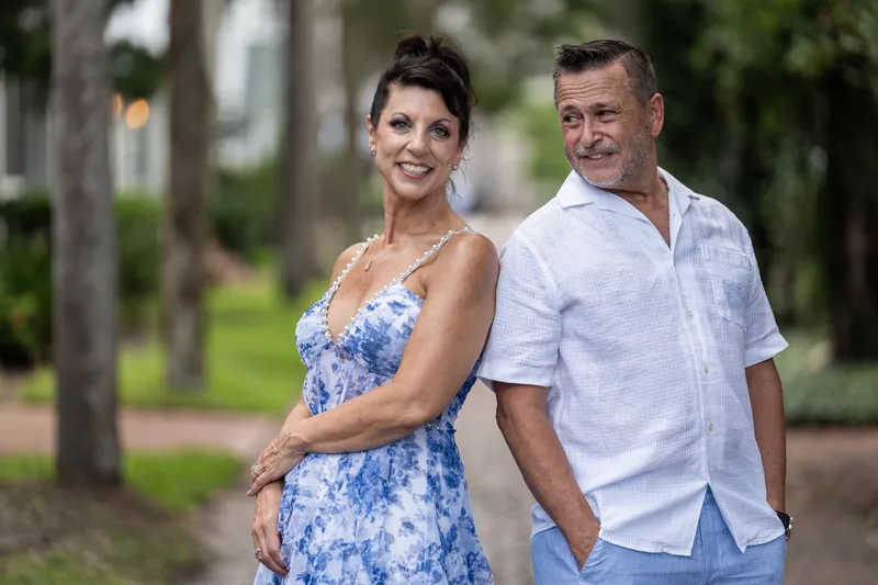 Mary in a blue floral dress and Brian in a light blue shirt walk arm-in-arm through a tree-shaded park setting.