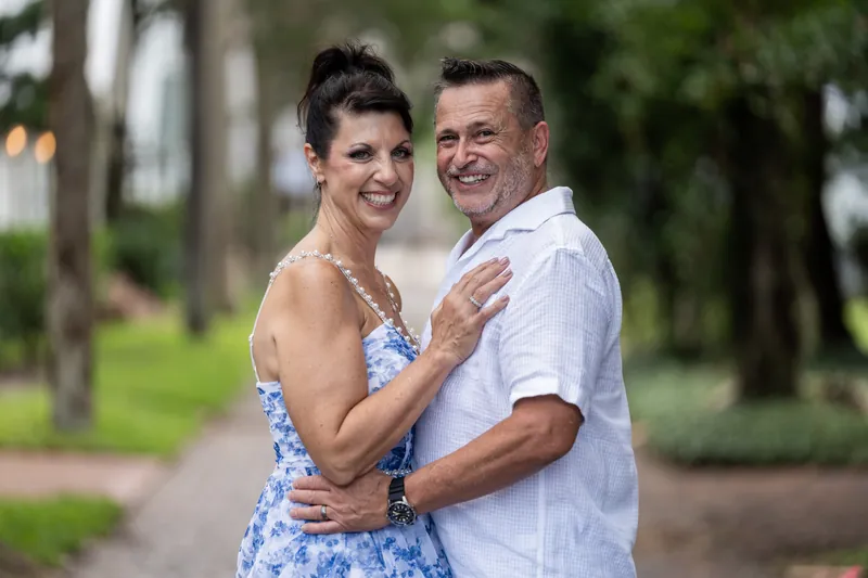 Mary and Brian embrace on a brick pathway with tall trees in the background, both smiling at the camera.