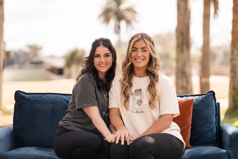 Bride and maid of honor laughing on blue couch in matching frog princess shirts