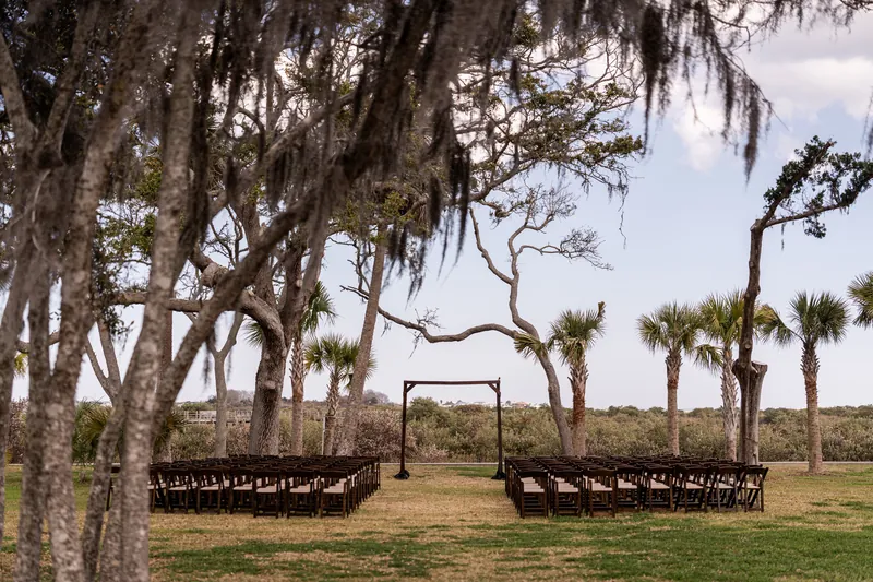 Ceremony setup with wooden chairs, arbor, palms, Spanish moss, and waterfront view