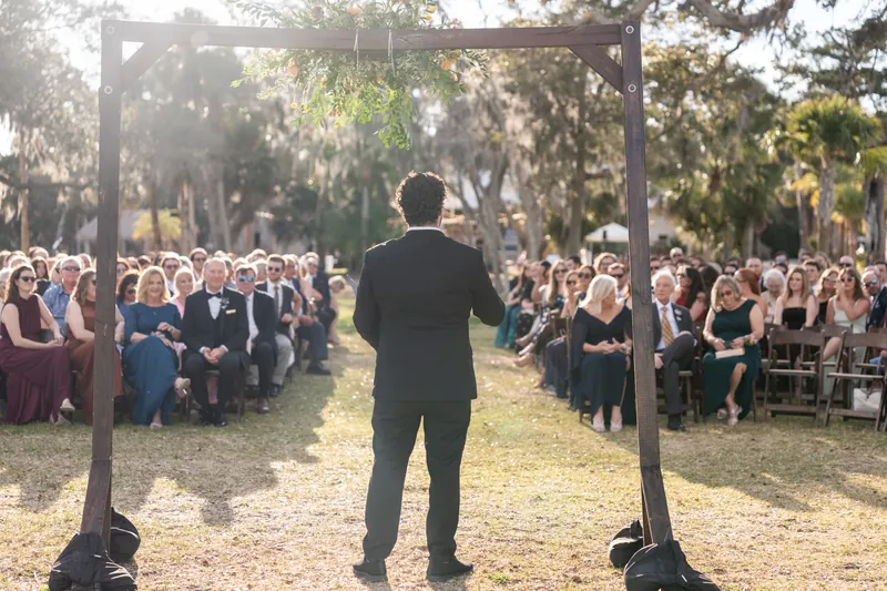 Groom standing at altar facing seated guests through wooden arbor