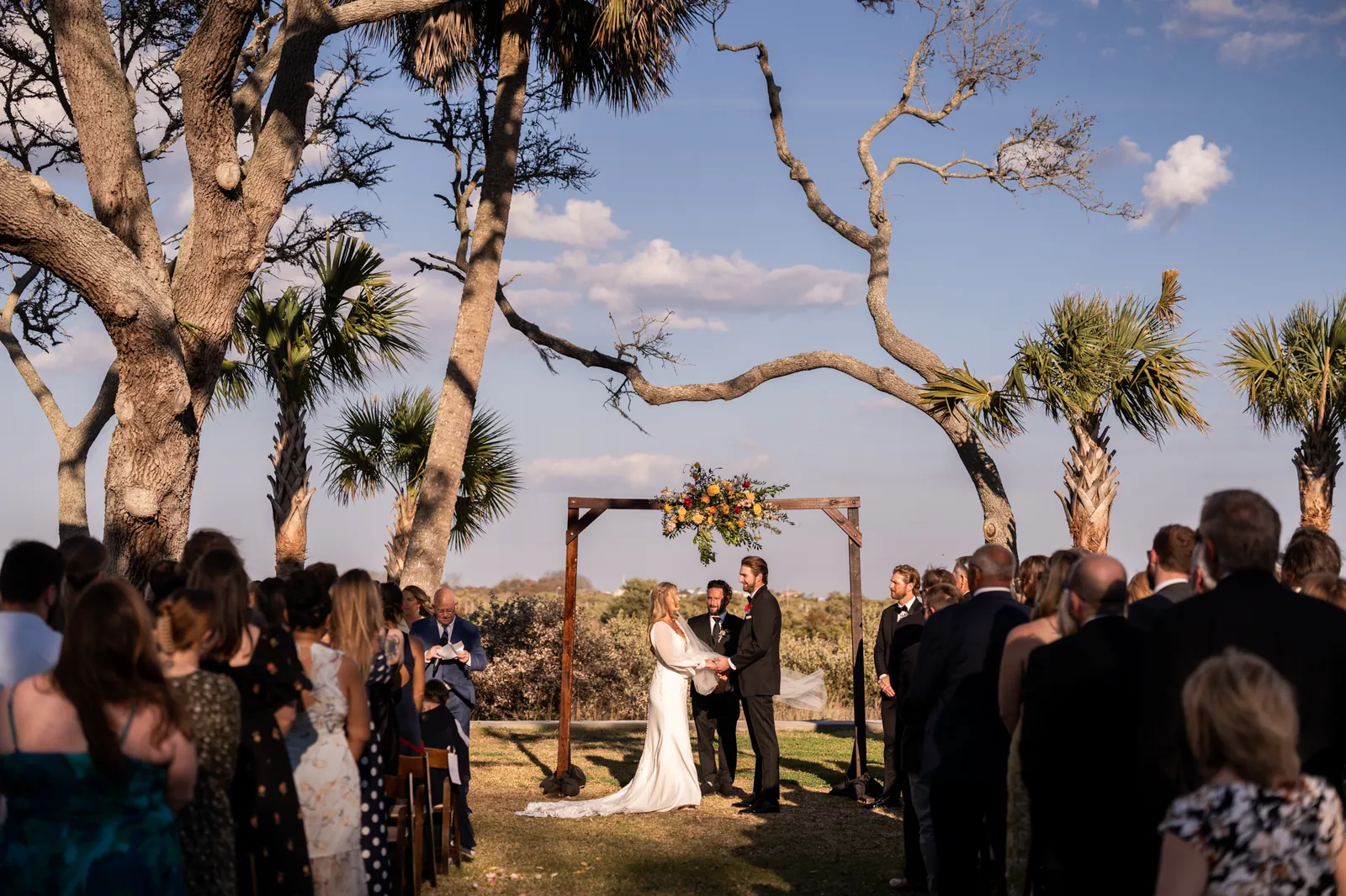 Guests stand beneath sprawling oaks with pale trunks as Mary and Gabe exchange vows beneath a wooden arch decorated