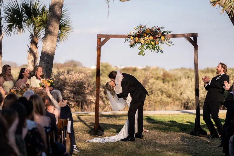 First kiss with groom dipping bride under arbor as wedding party cheers