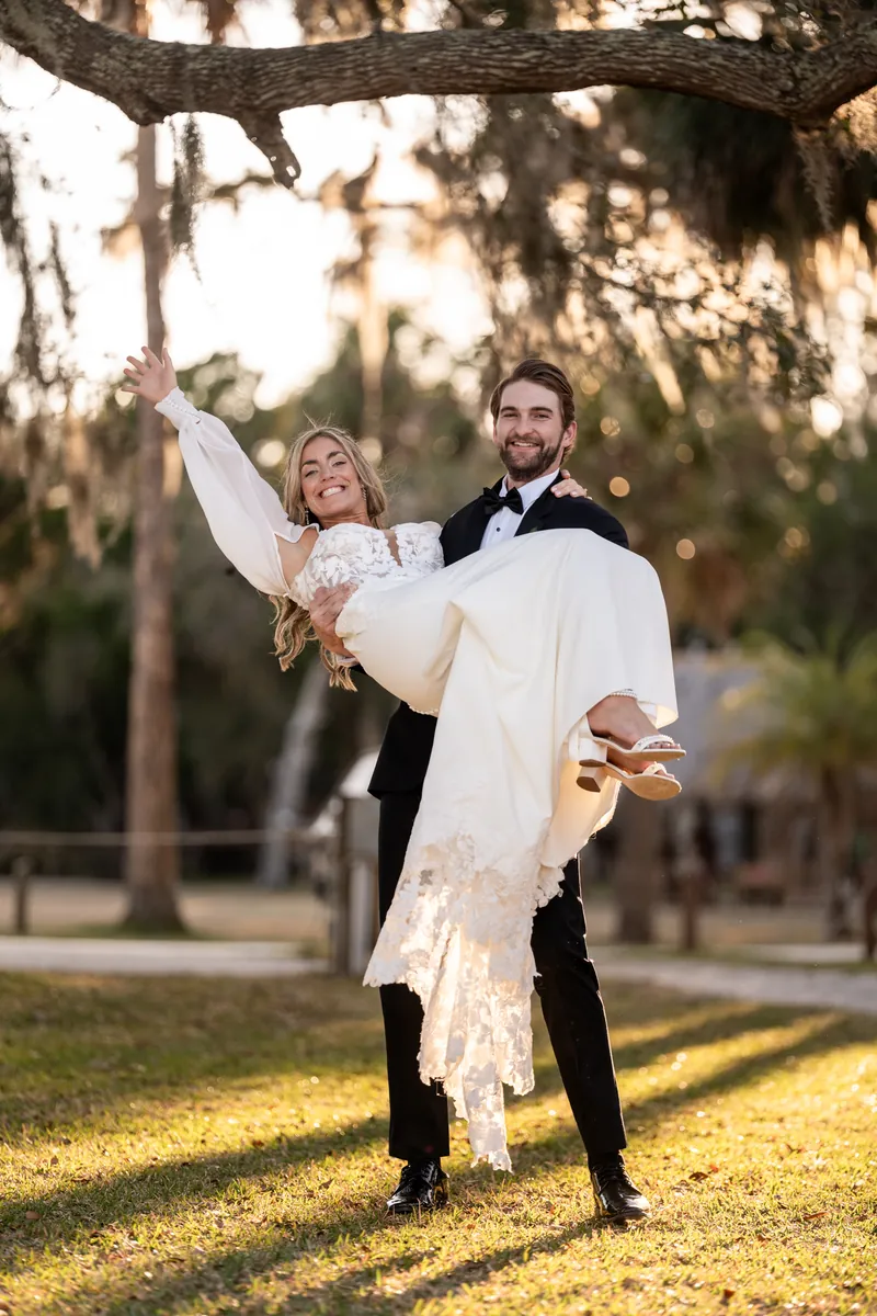 Groom carrying bride under oak tree branch in golden hour light