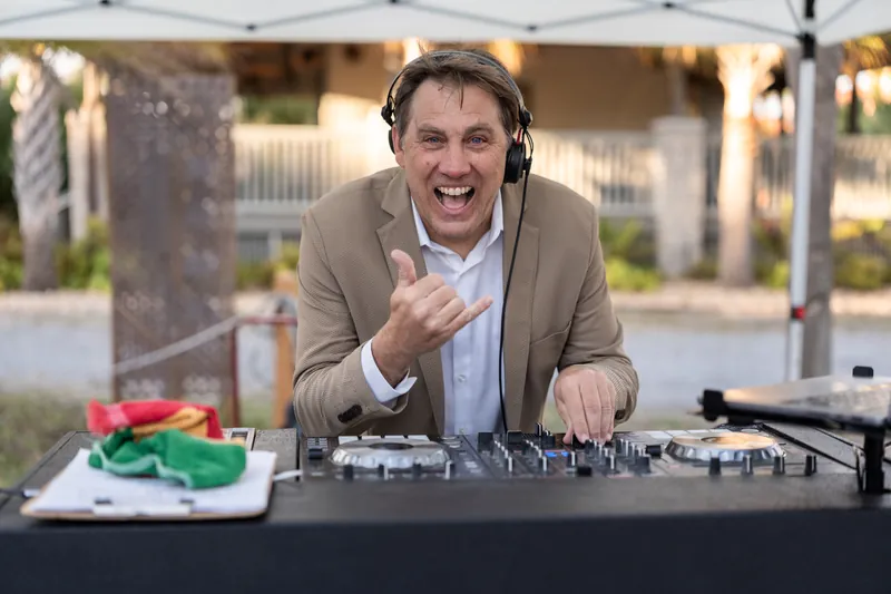 DJ giving shaka sign at turntables wearing tan suit and headphones