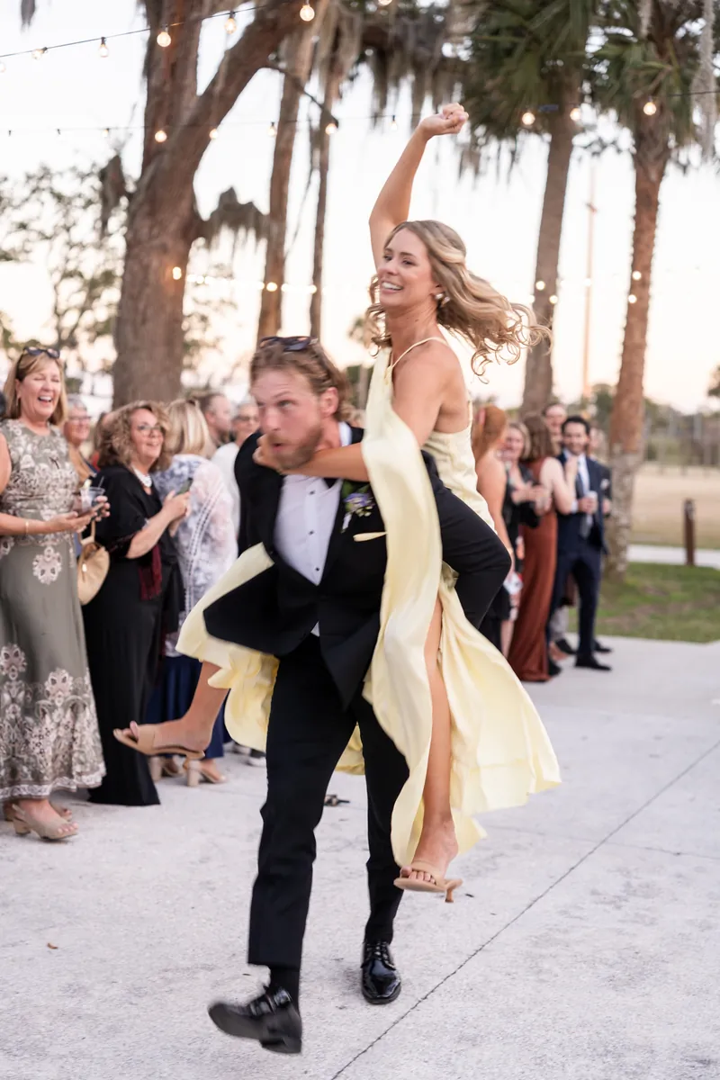 Groomsman piggyback riding bridesmaid in yellow dress during bridal party entrance