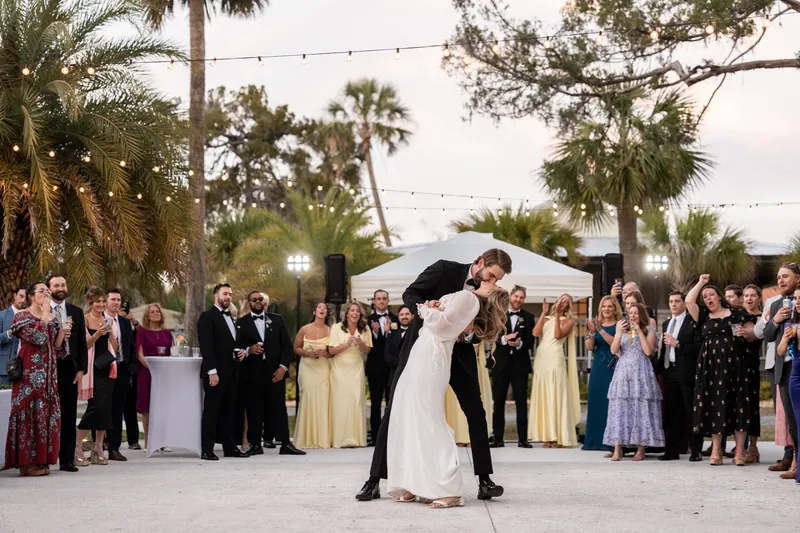 First dance dip kiss under string lights with full wedding party watching