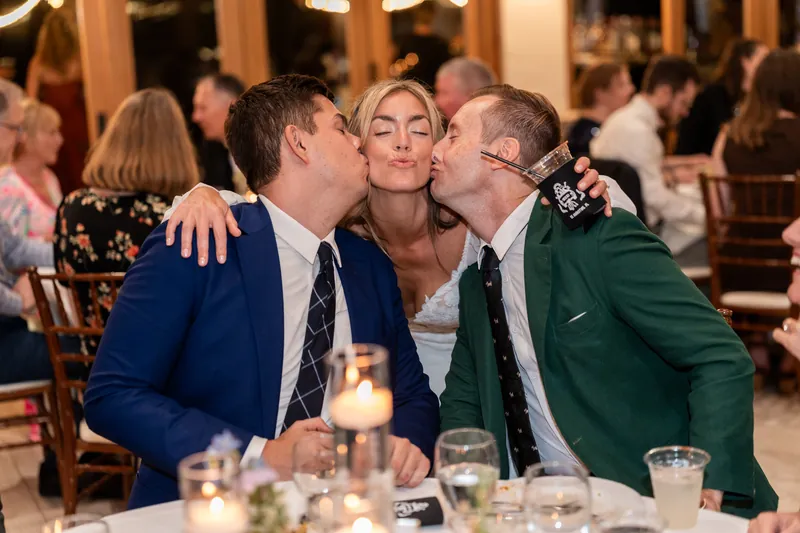 Bride getting kissed on both cheeks by two friends at reception table