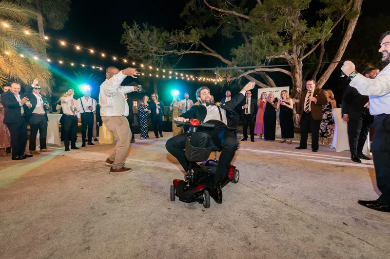 Groom riding mobility scooter across dance floor as guests cheer