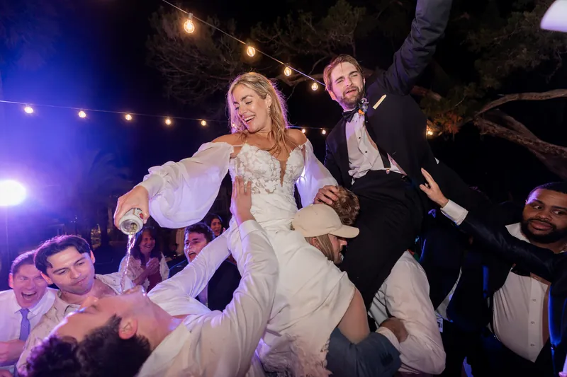 Couple lifted by guests as bride pours champagne under purple uplighting