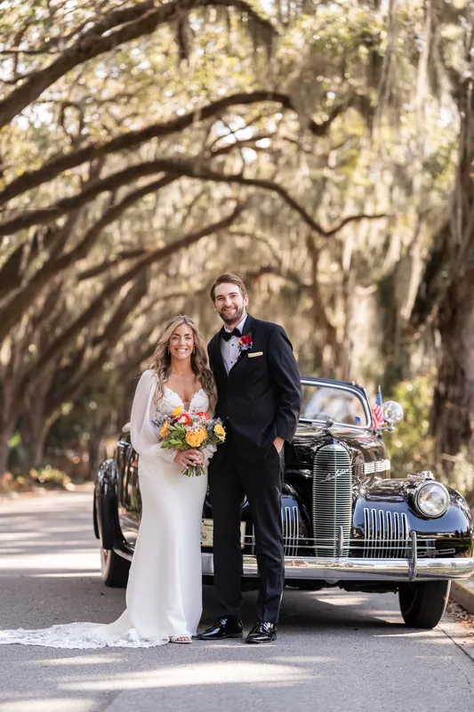Mary holds a colorful bouquet beside Gabe near a vintage blue car beneath moss-draped oak trees on Magnolia Avenue.