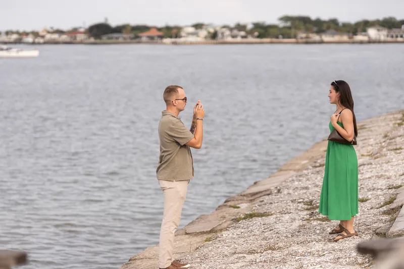 Mason kneels on a rocky beach in a tan suit while Caitlin stands in a green dress across from him, hand raised to her mouth in surprise.