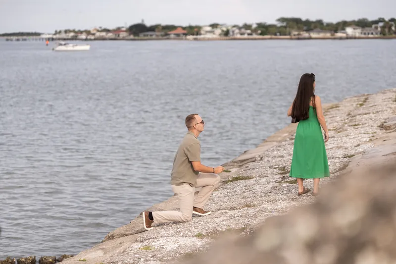 Mason on one knee on the beach rocks, facing Caitlin in her green dress as the bay and shoreline stretch behind them.