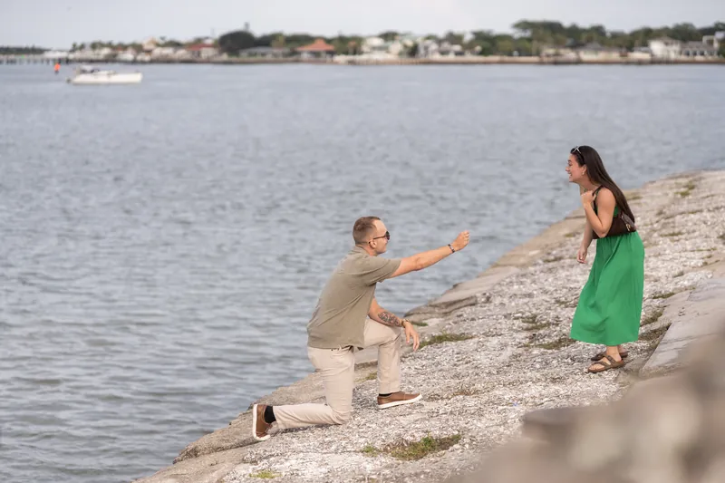 Mason reaches toward Caitlin on the beach, both positioned on the rocky ground with water and houses visible across the bay.