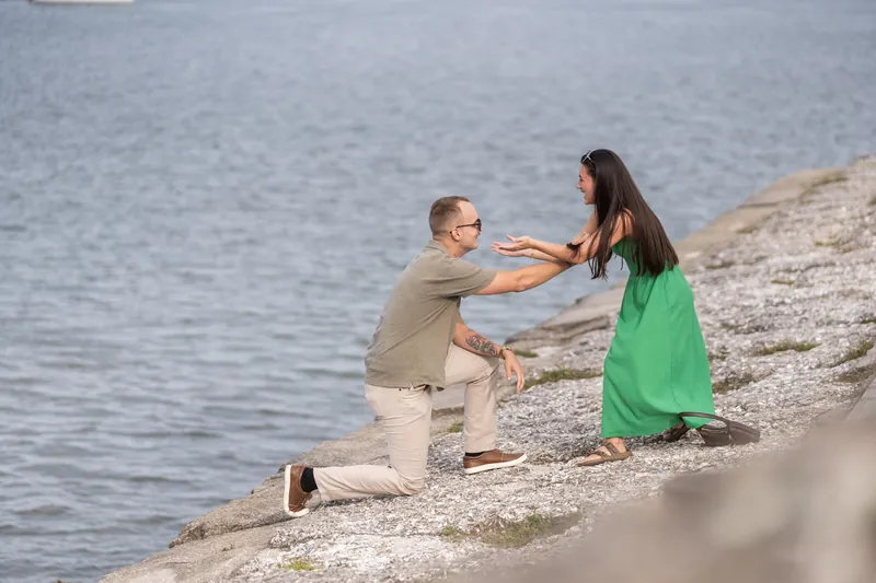 Mason kneels and extends his hands toward Caitlin, who stands in green facing him on the pebbly beach.