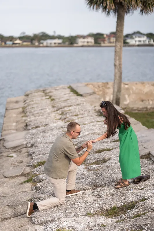 Mason kneels beside a palm tree trunk on the beach while Caitlin stands facing him in her green dress.