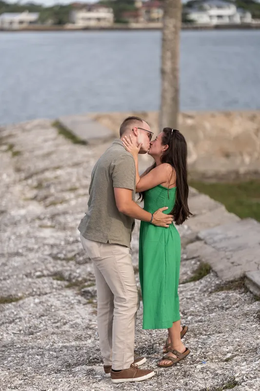 Mason and Caitlin embrace and kiss on the rocky beach, a palm tree and waterfront homes visible behind them.