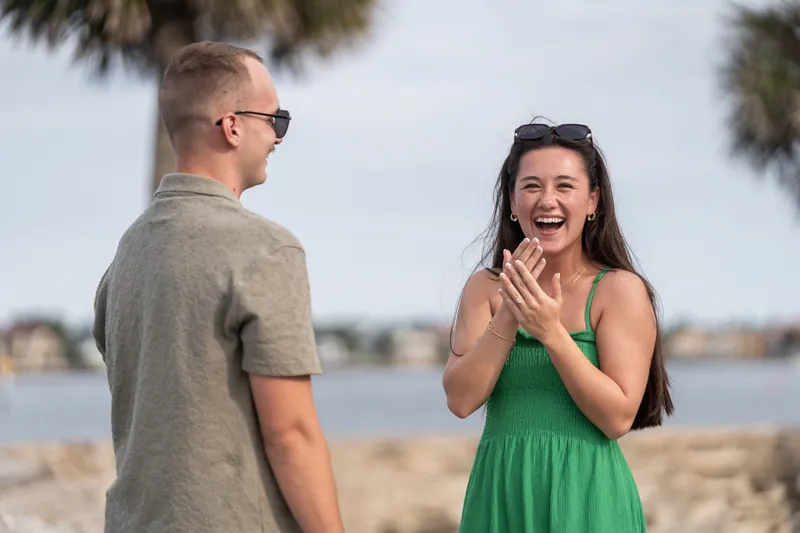 Woman in a green dress laughs with hand near her face as man in gray shirt stands beside her by the water.